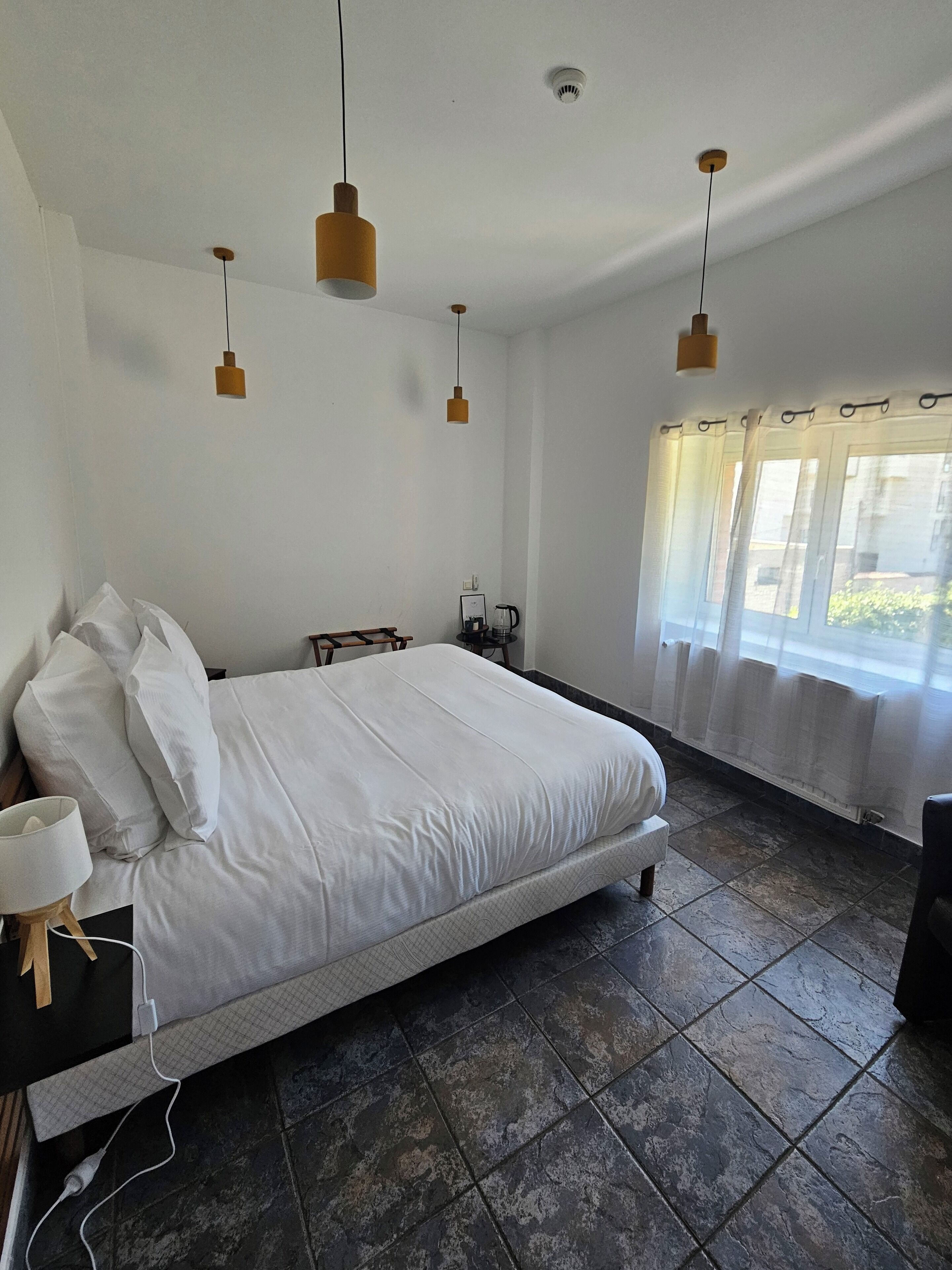 A bedroom with a large bed, a bedside table with a lamp, and a window with sheer curtains.