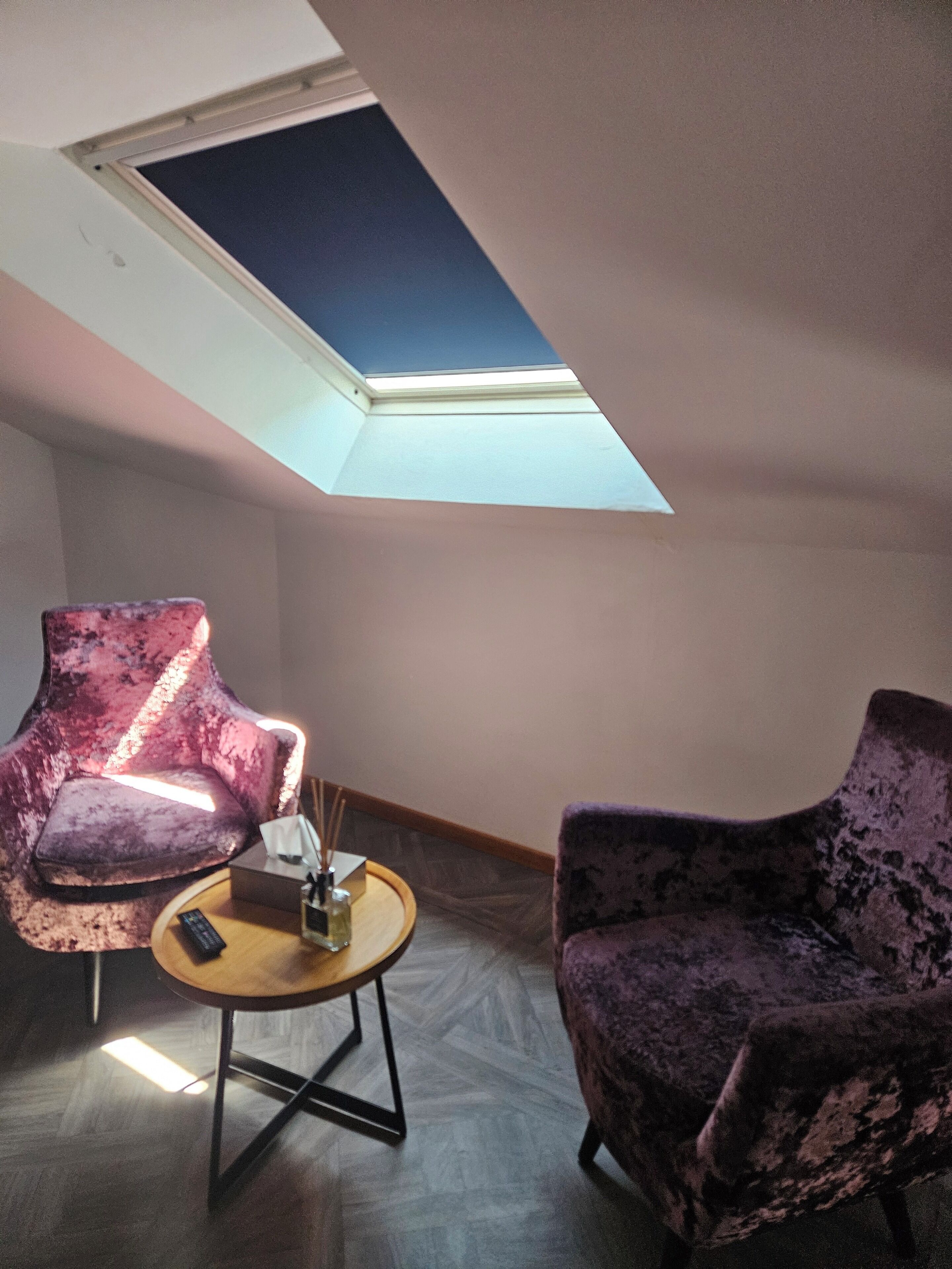 Une pièce dotée d’une lucarne, de deux fauteuils violets, d’une petite table ronde sur laquelle se trouvent une télécommande et une bouteille, et d’un plancher en bois.