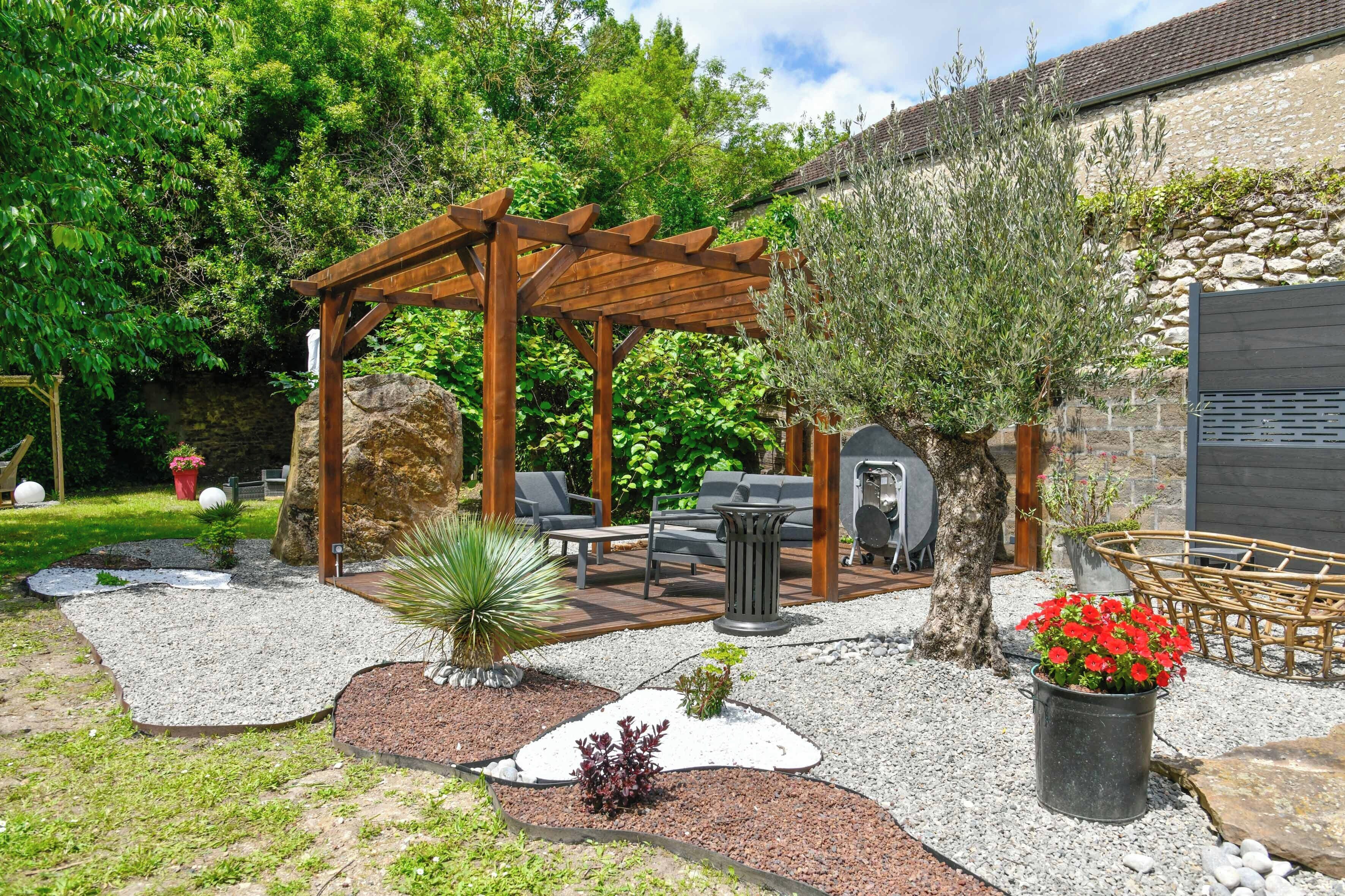 Une pergola en bois avec des sièges, un mur en pierre et un jardin avec des allées de gravier et des plantes.