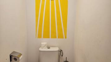Une petite salle de bain comprenant des toilettes, un rouleau de papier toilette, une poubelle et un élément décoratif fixé au mur au-dessus des toilettes.