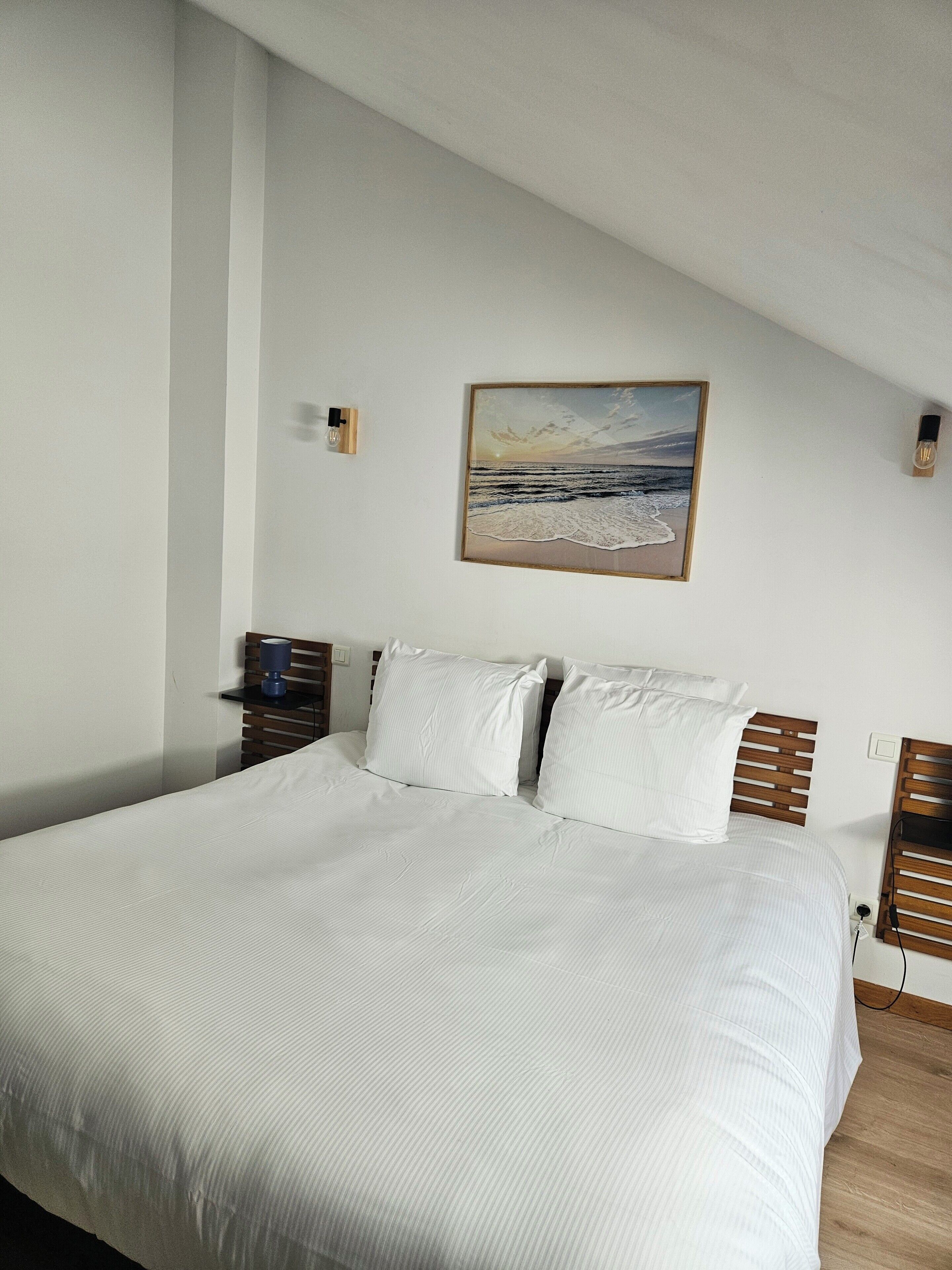 Cama bem arrumada com lençóis brancos, cabeceira de madeira e uma pintura de uma praia na parede acima.