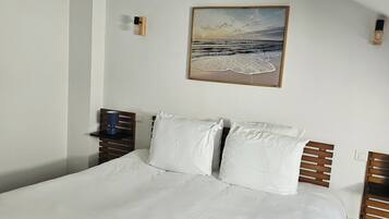 Cama bem arrumada com lençóis brancos, cabeceira de madeira e uma pintura de uma praia na parede acima.