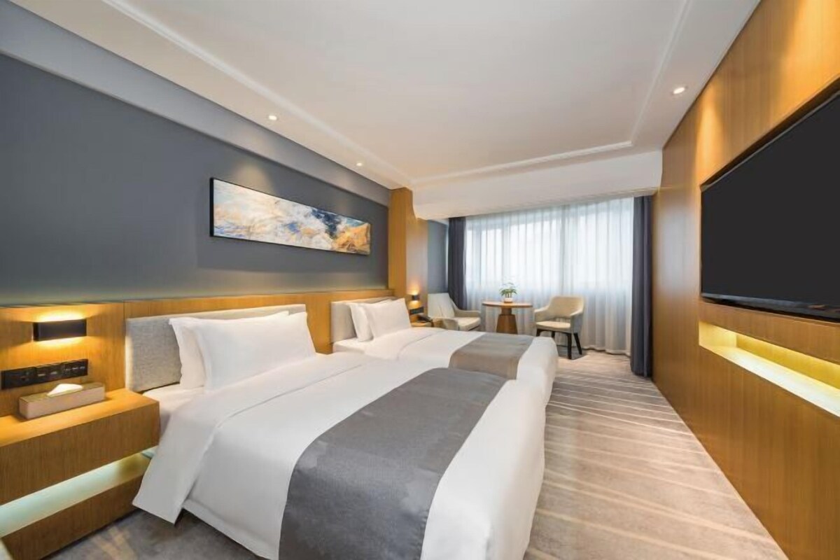 A modern hotel room with a large bed, bedside tables, a flat-screen TV, a small table with chairs, and a large window with curtains.