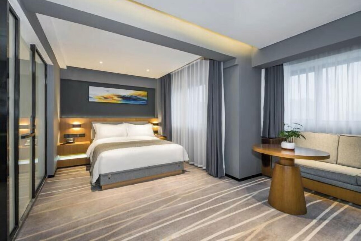 A modern hotel room with a large bed, bedside tables, a sofa, and a view of the outdoors.