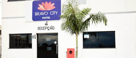 Entrada de un hotel con una recepción, un letrero del Hotel Bravo City y una palmera en primer plano.