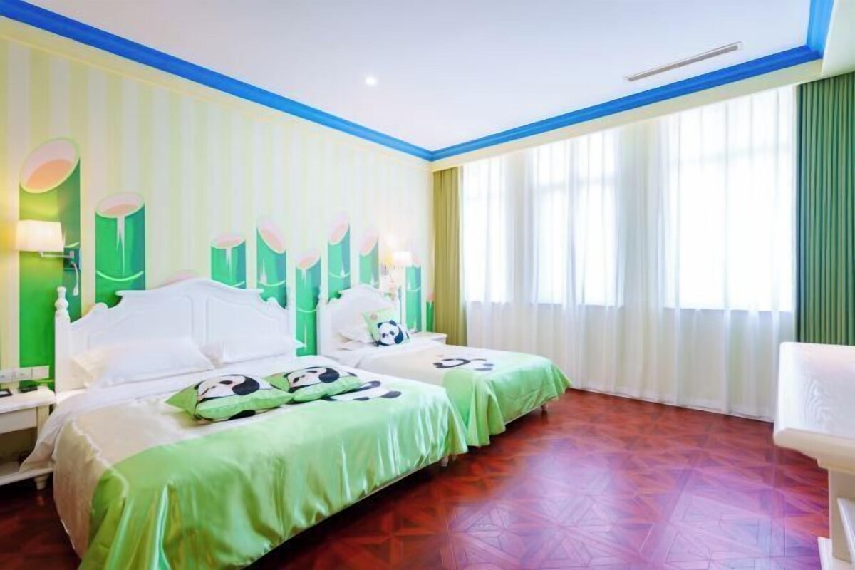 A hotel room with two beds, green bedding, a white headboard, a nightstand, a lamp, and a window with sheer curtains.