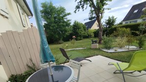 Un patio con una mesa redonda y sillas, una sombrilla azul, y un jardín con una estatua y árboles.