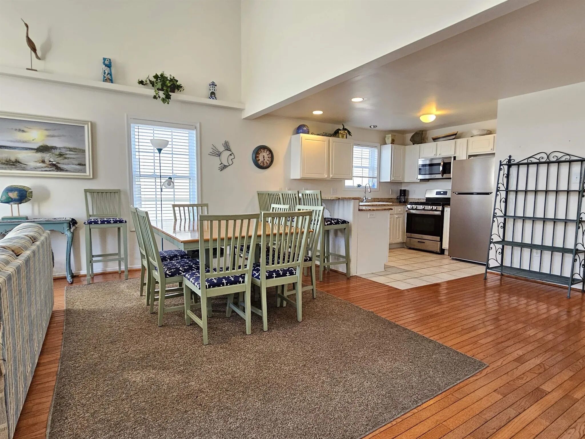 A spacious kitchen with wooden flooring, white cabinets, a central island, and a dining area with green chairs and patterned cushions.