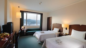 Une chambre d’hôtel avec deux lits, un bureau avec une télévision, une lampe de bureau, une fenêtre donnant sur la mer et une petite table agrémentée de fleurs.