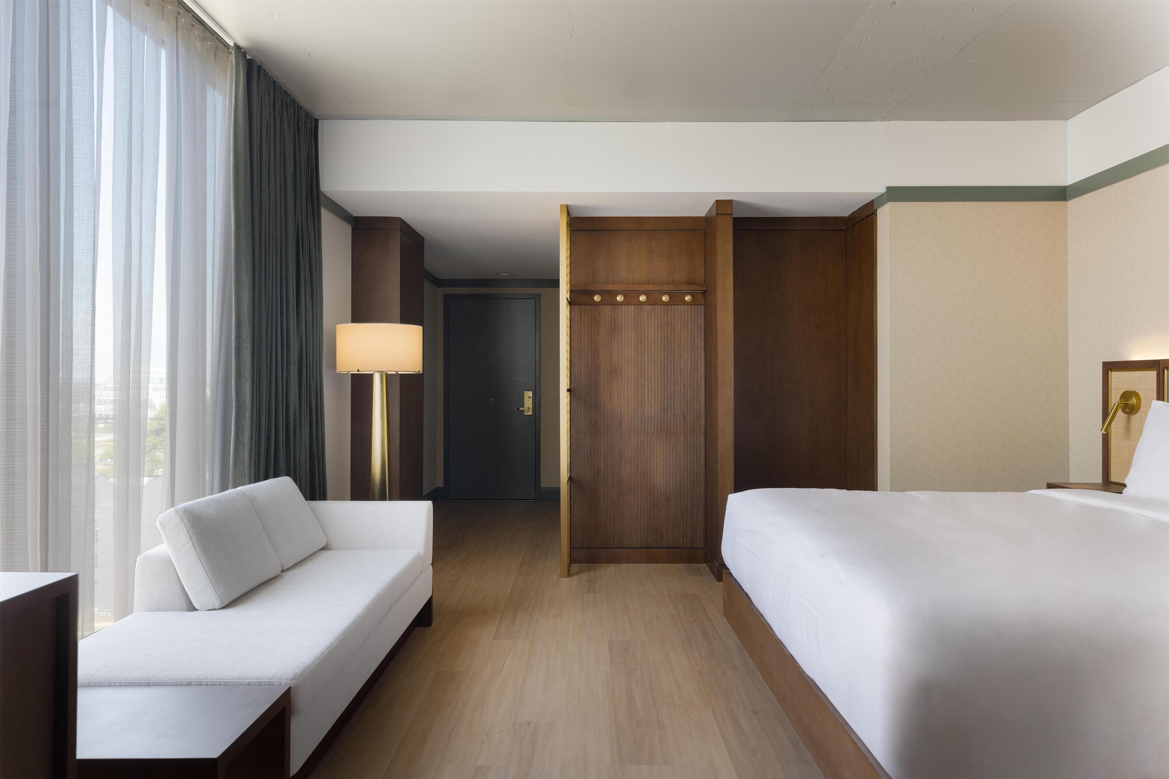 Une chambre d’hôtel avec un canapé, un lit, une table de chevet, une lampe et une vue sur l’extérieur par la fenêtre.