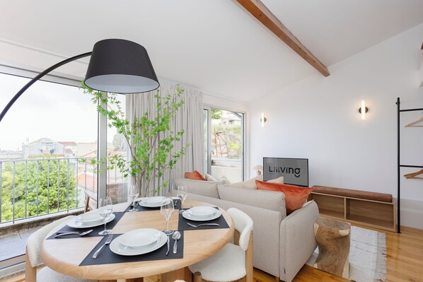 A modern living room with a dining area, a sofa, a bench, and a large window with a view of greenery and buildings. - Liiiving - Mezzanine Delight Balcony (Porto)