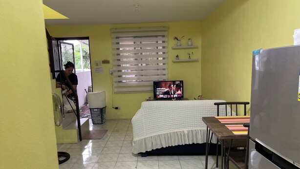 A small, yellow-walled room with a bed, a TV, a refrigerator, and a window with blinds.