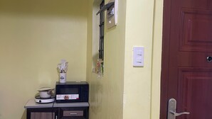 A small kitchen with a microwave, a kettle, and a cabinet. There is a wooden door and a light switch on the wall.