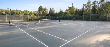 Un court de tennis en plein air, doté d’un filet et entouré d’arbres et d’une clôture.