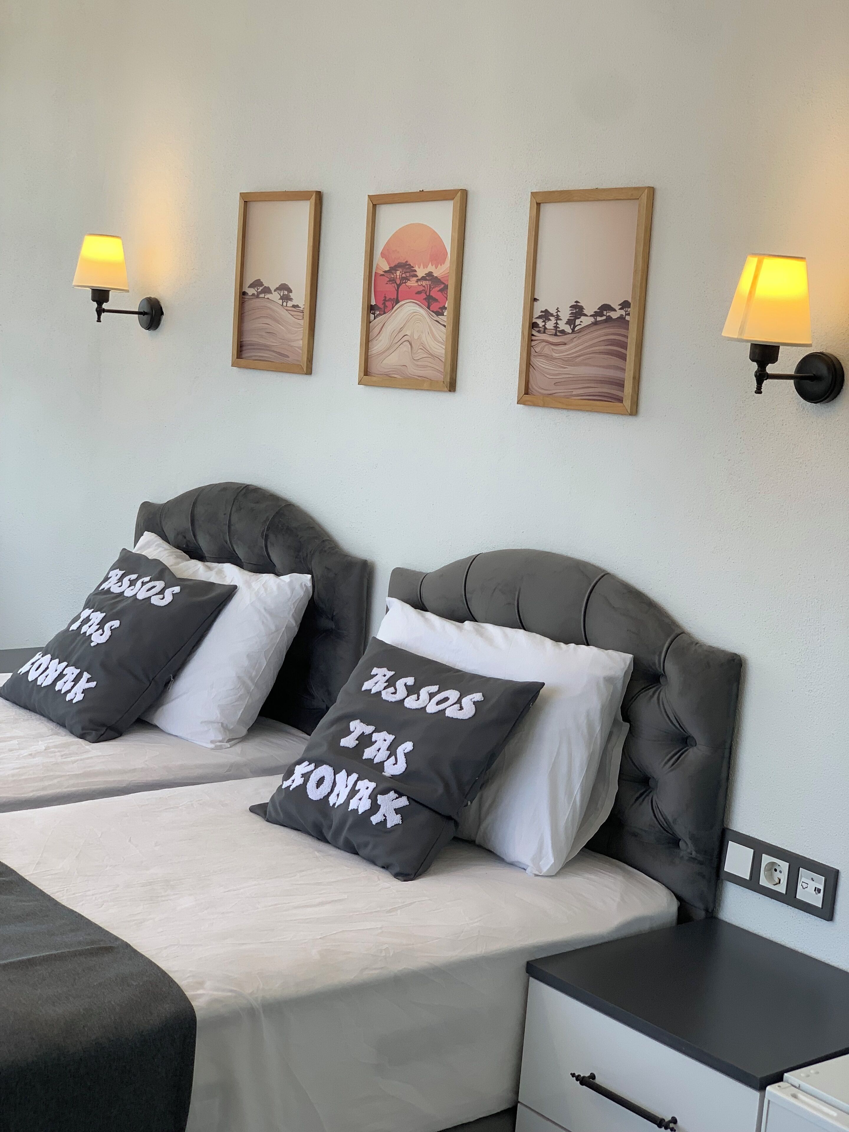 Ein Hotelzimmer mit zwei Betten, jeweils mit einem gesteppten Kopfteil und einem schwarzen Kopfkissen mit weißer Schrift. Darüber an der Wand hängen drei gerahmte Kunstwerke, und je eine Wandleuchte befindet sich an beiden Seiten.