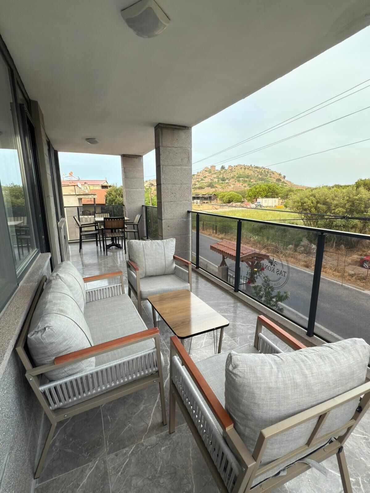 A balcony with a sofa, chairs, and a table, overlooking a road and greenery.