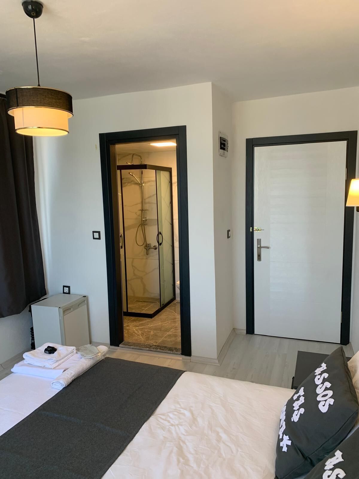 A hotel room with a bed, a bedside table, a small desk, a shower area, and a door.