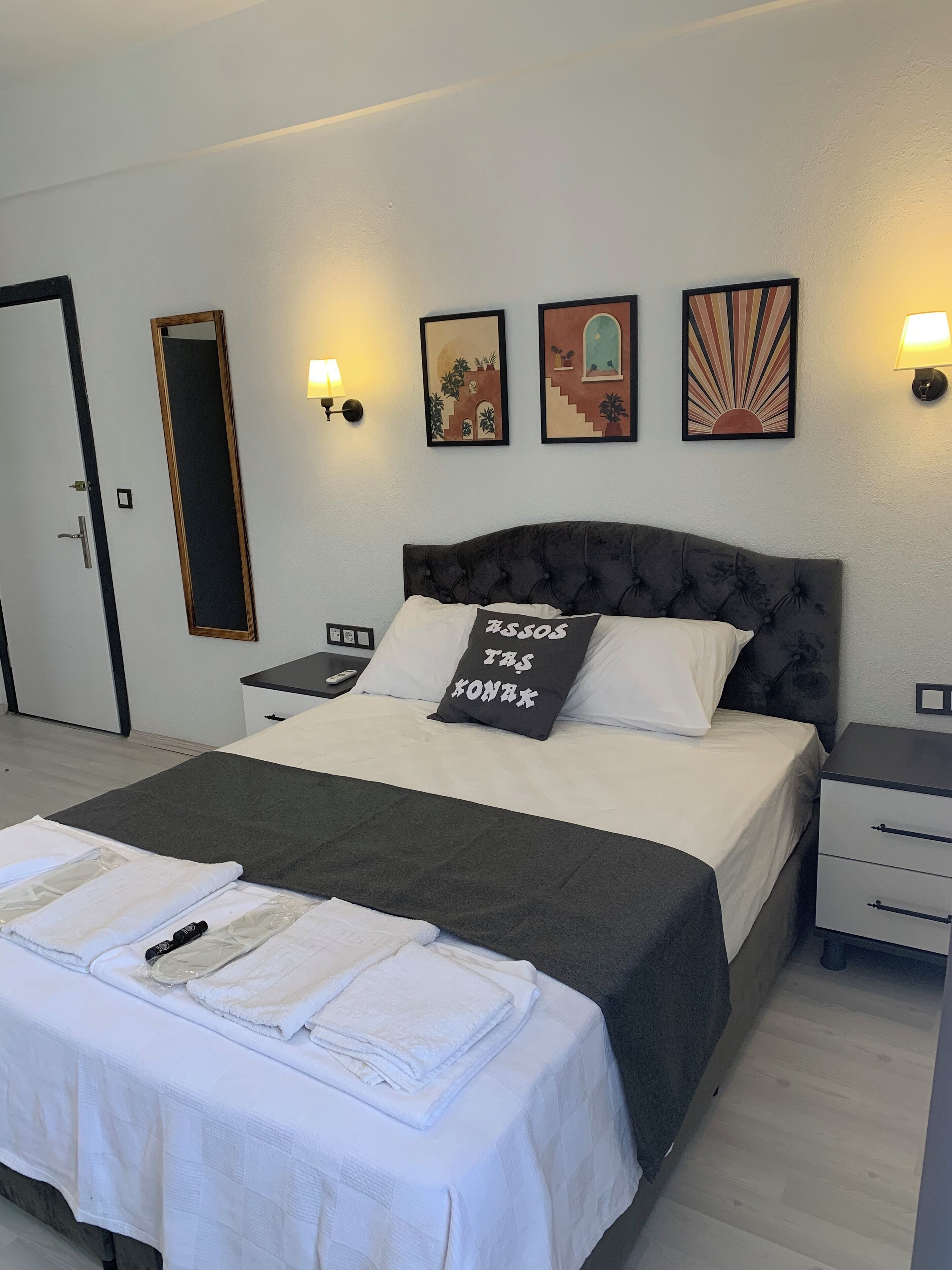 A neatly made bed with a grey headboard, white bedding, and a pillow with text. There are wall-mounted lamps, a mirror, and three framed artworks above the bed.
