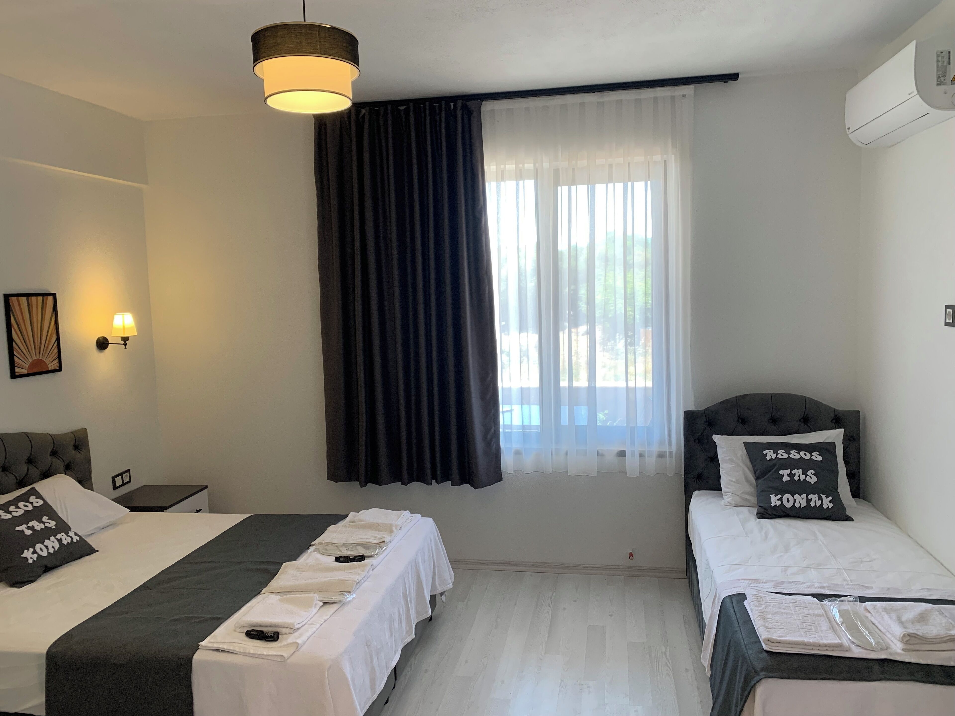 A hotel room with two beds, a large window with sheer curtains, a wall-mounted air conditioner, and a ceiling light fixture.