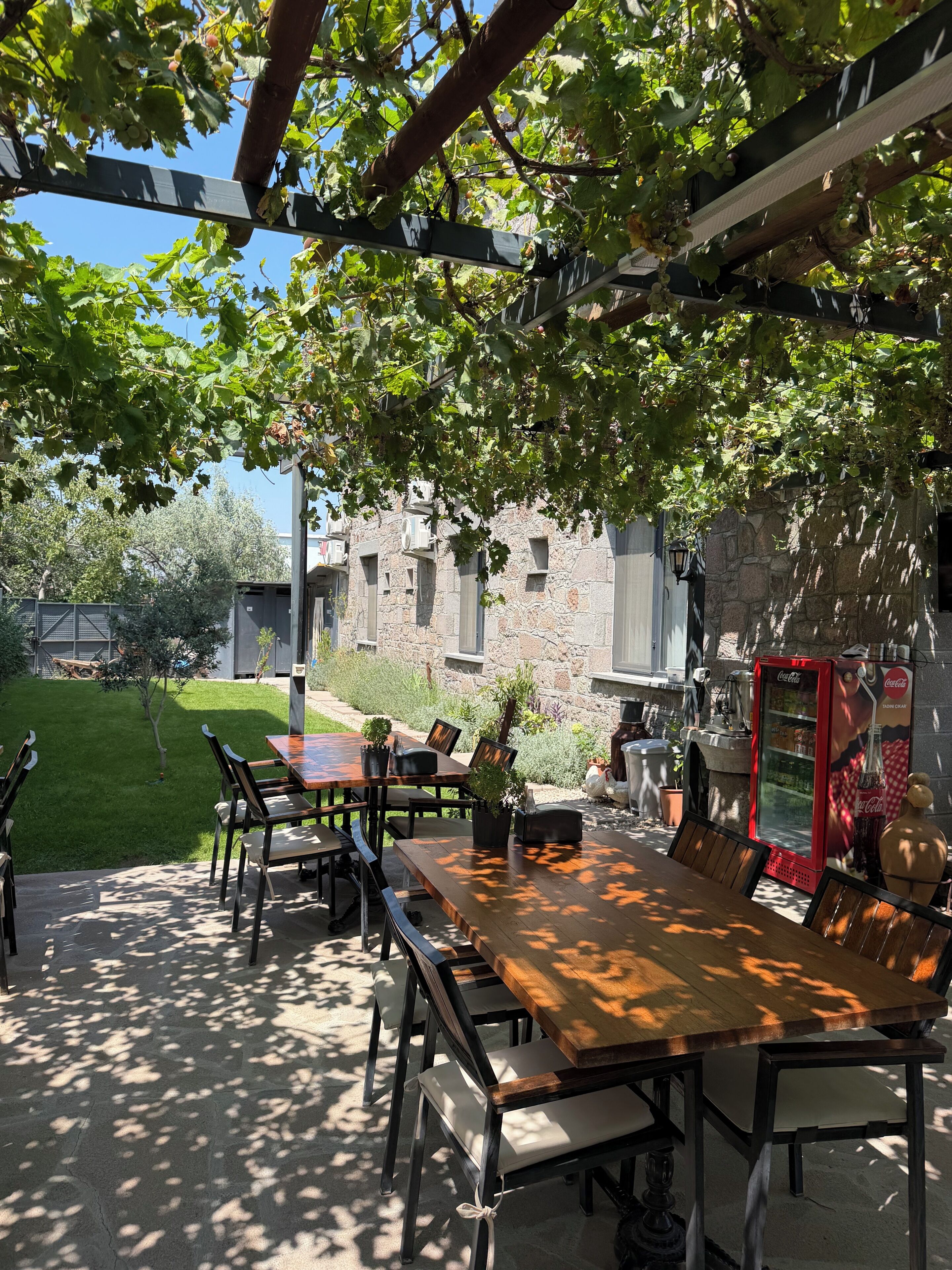 An outdoor patio with a wooden table and chairs, a stone building in the background, and a Coca-Cola vending machine.