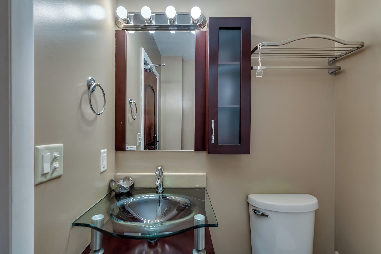 Un baño con un lavabo de vidrio, un espejo con iluminación, un perchero para toallas y un inodoro.