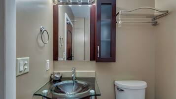 Un baño con un lavabo de vidrio, un espejo con iluminación, un perchero para toallas y un inodoro.