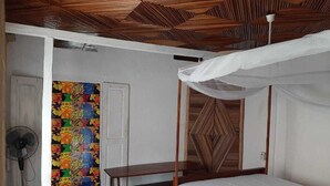 A bedroom with a wooden ceiling, a bed with a white canopy, a colorful door, and a fan.
