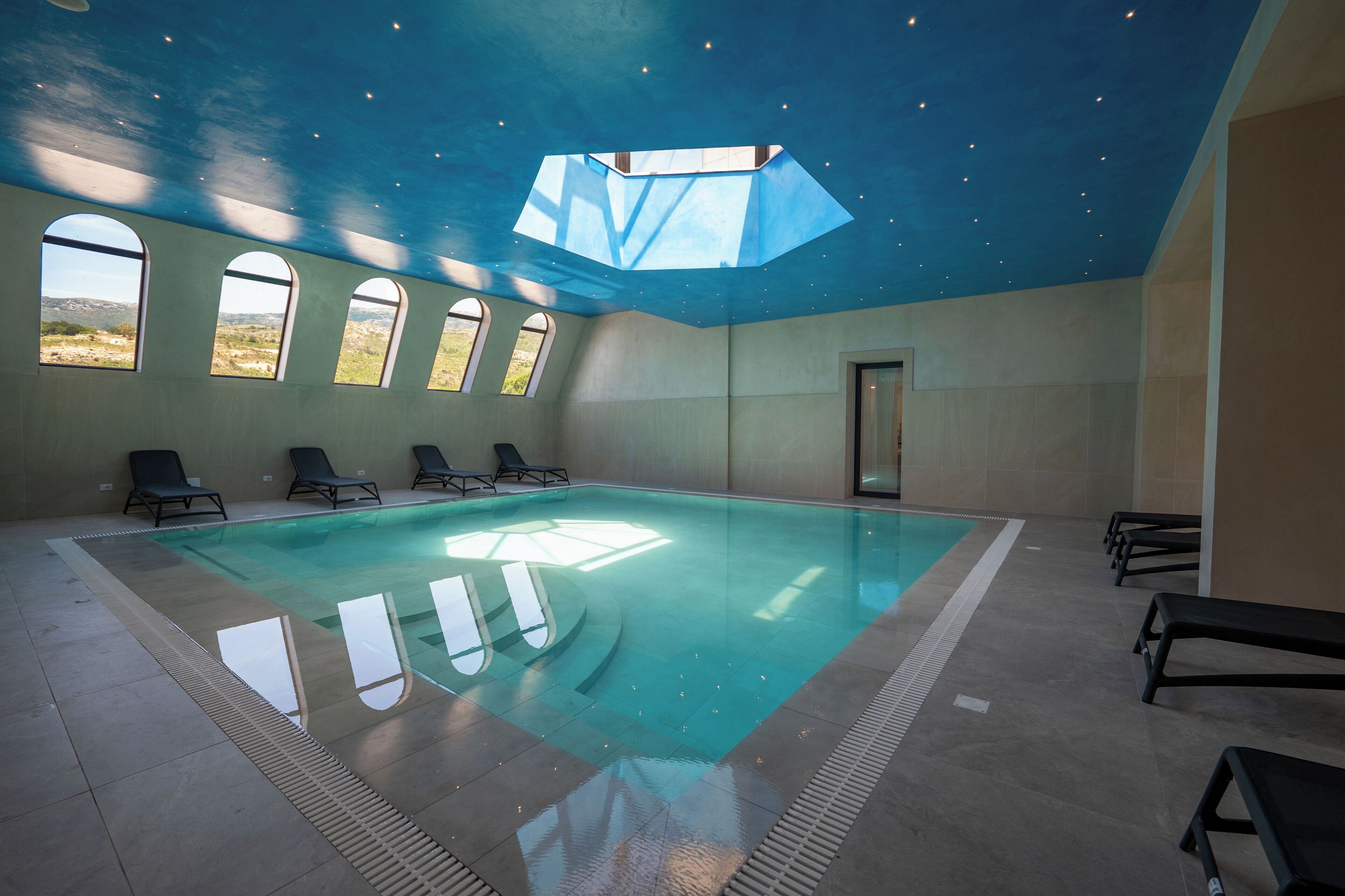 An indoor swimming pool with clear glass walls, a geometric ceiling design, and multiple windows offering views of the outdoors.