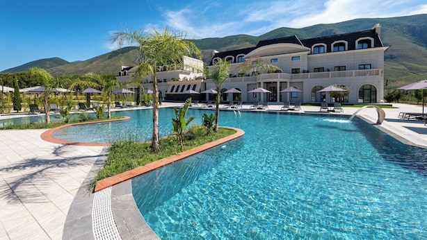 A large swimming pool with a curved edge, surrounded by lounge chairs and umbrellas, with a building and mountains in the background. - Acros Resort (Orikum)