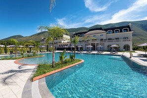 A large swimming pool with a curved edge, surrounded by lounge chairs and umbrellas, with a building and mountains in the background. - Acros Resort (Orikum)