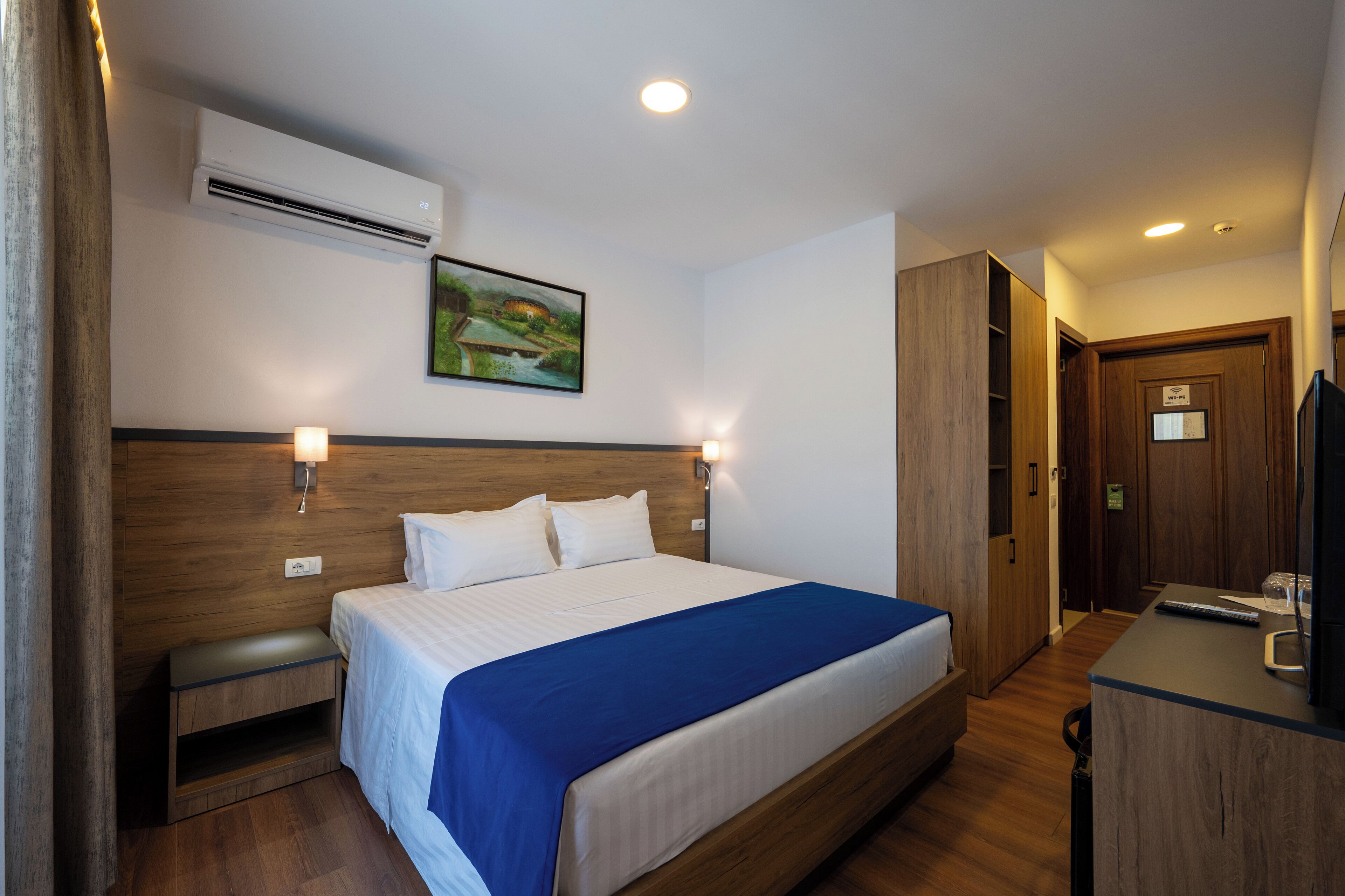 A hotel room with a wooden bed, a desk with a computer, an air conditioner, and a painting on the wall.