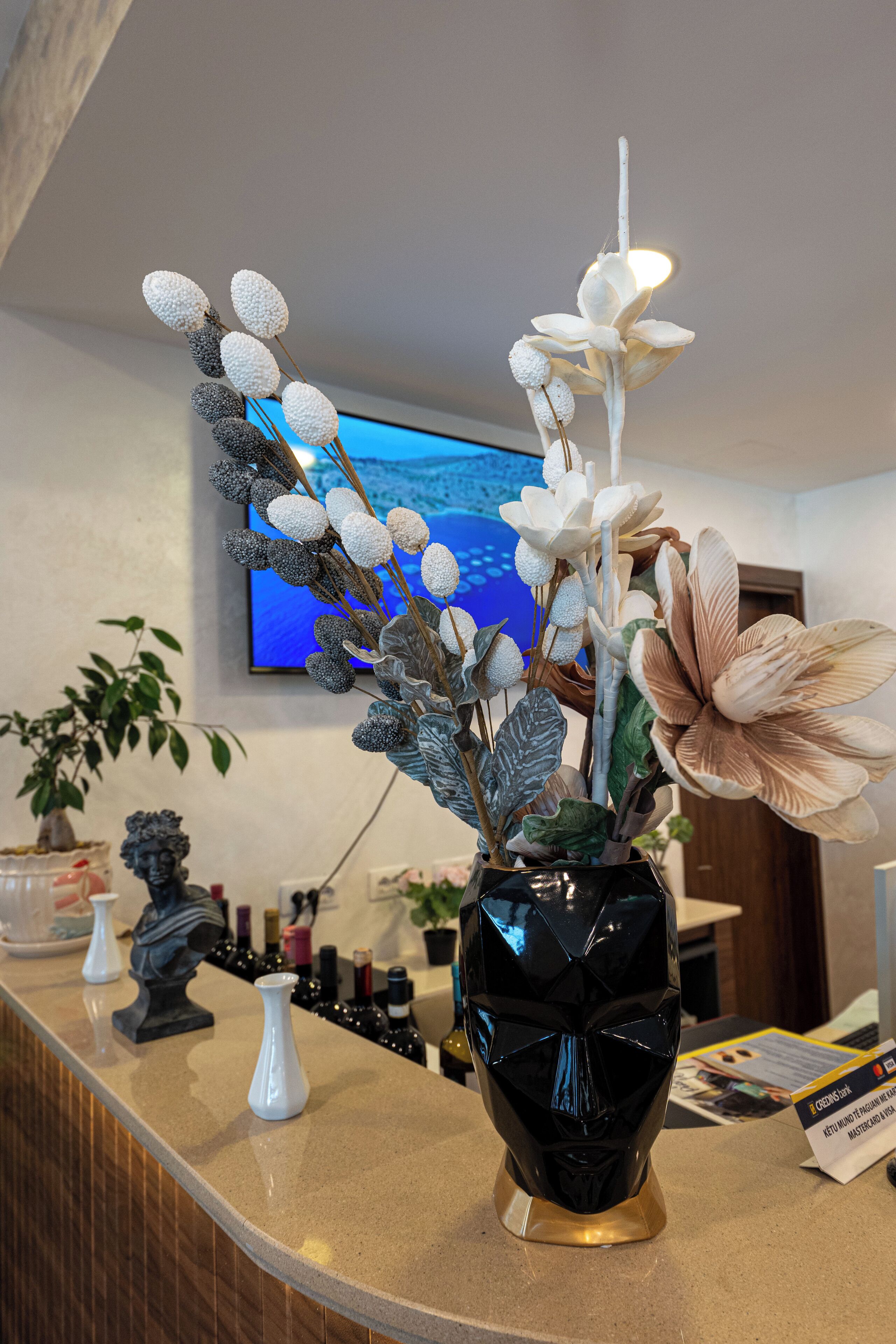 A reception area with a counter, a black vase with flowers, a television, and a statue.