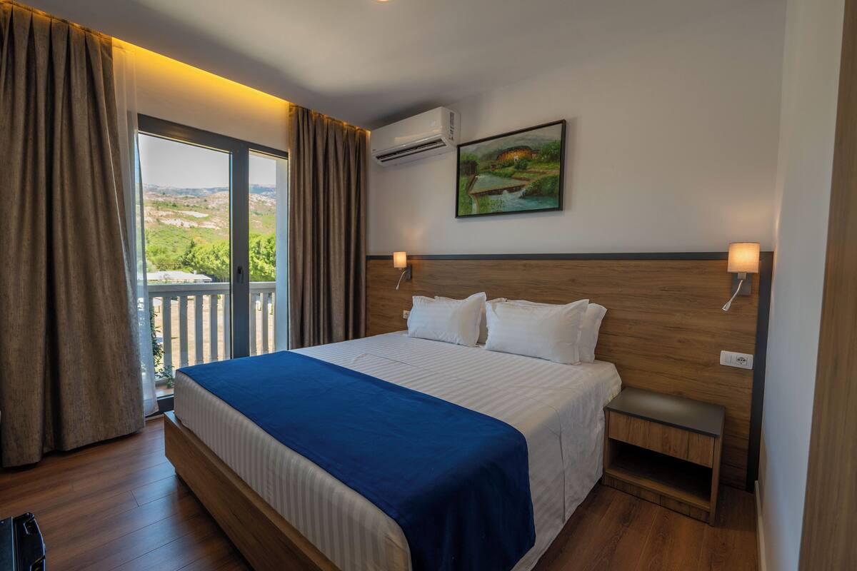 Habitación de hotel con una cama grande, cabecera de madera, mesita de noche, unidad de aire acondicionado, un cuadro en la pared y vistas por la ventana.