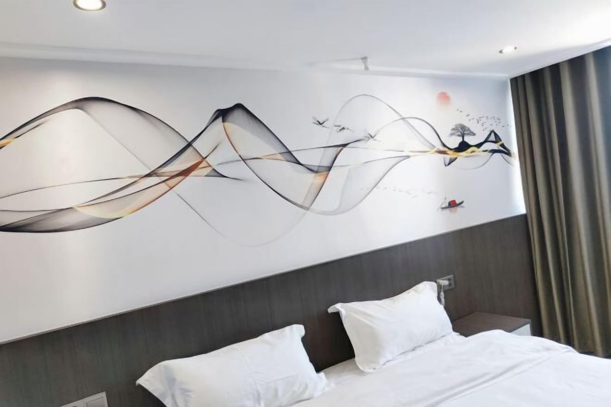 Quarto de hotel moderno com um grande mural abstrato na parede, uma cama bem arrumada com roupa de cama branca e cabeceira escura.