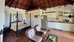 A bedroom with a four-poster bed, a sitting area with wicker furniture, and a kitchen with white cabinets and a tiled backsplash.