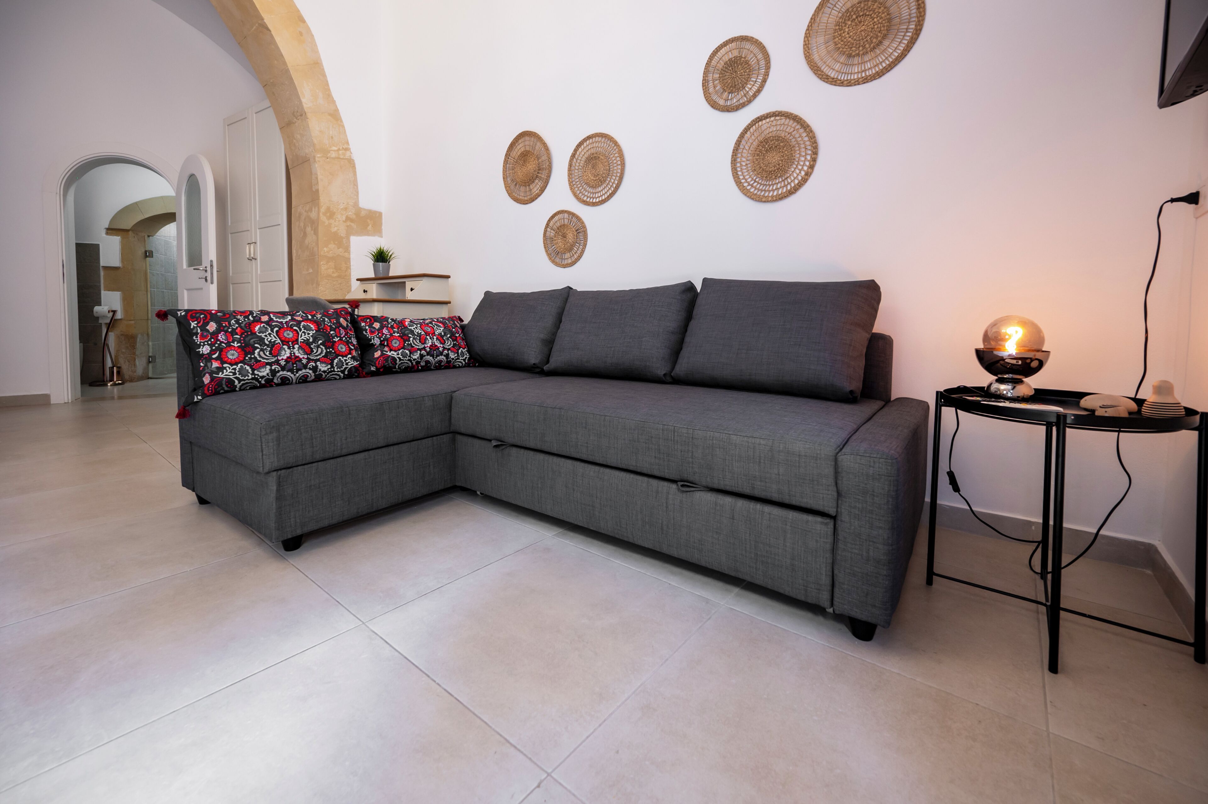 Una sala de estar moderna con un sofá seccional gris, decoración artística de tejido en la pared y una mesita con una lámpara.