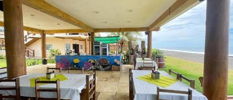 An outdoor dining area with tables and chairs, a mural, and a view of the beach.