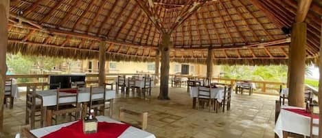 Un área de comedor al aire libre con mesas y sillas de madera, techo de paja y piso de baldosas.