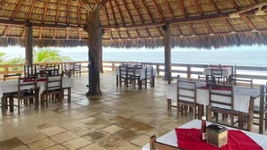 Un área de comedor al aire libre con mesas y sillas, techo de paja y vista al océano.