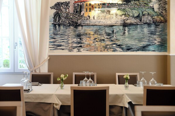 A dining area with a table set for four, a large painting of a coastal scene above, and a window with curtains. - Hotel Ristorante La Vignetta Cernobbio (Cernobbio)