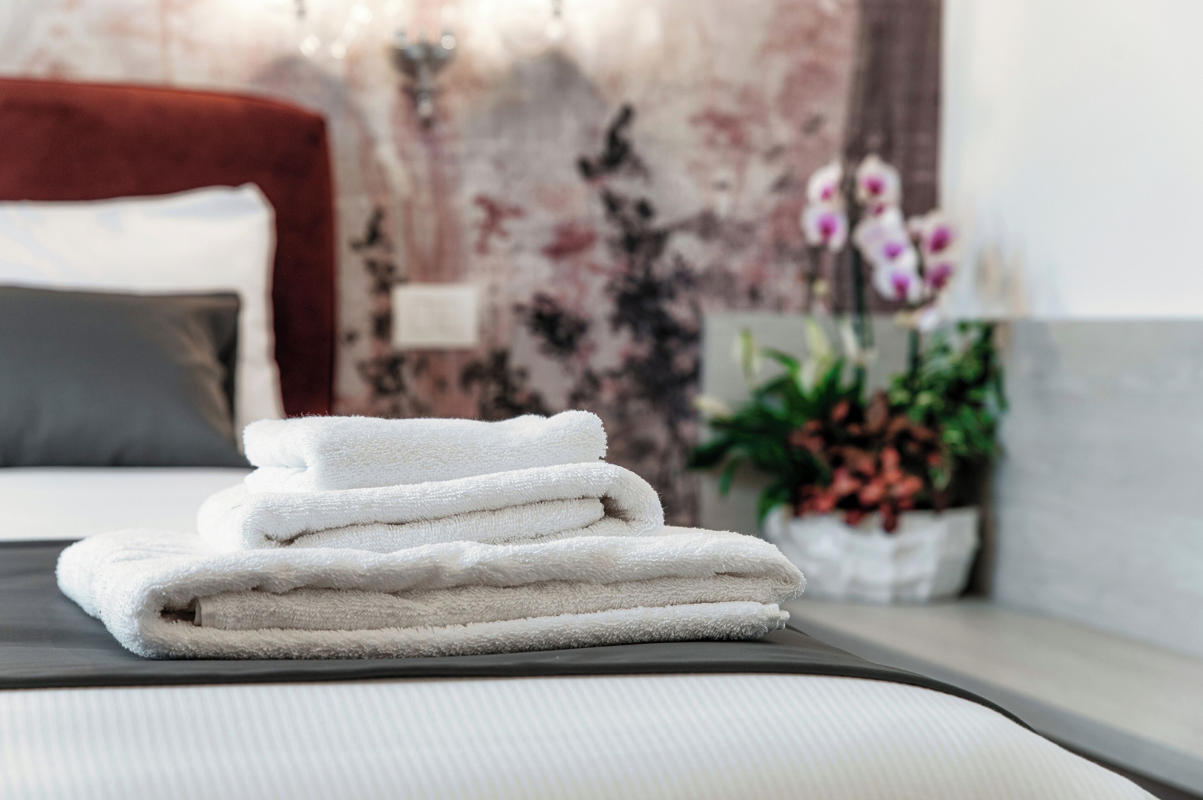 Een netjes opgevouwen stapel witte handdoeken op een bed met een donker hoofdbord en een bloemendecor op de muur.
