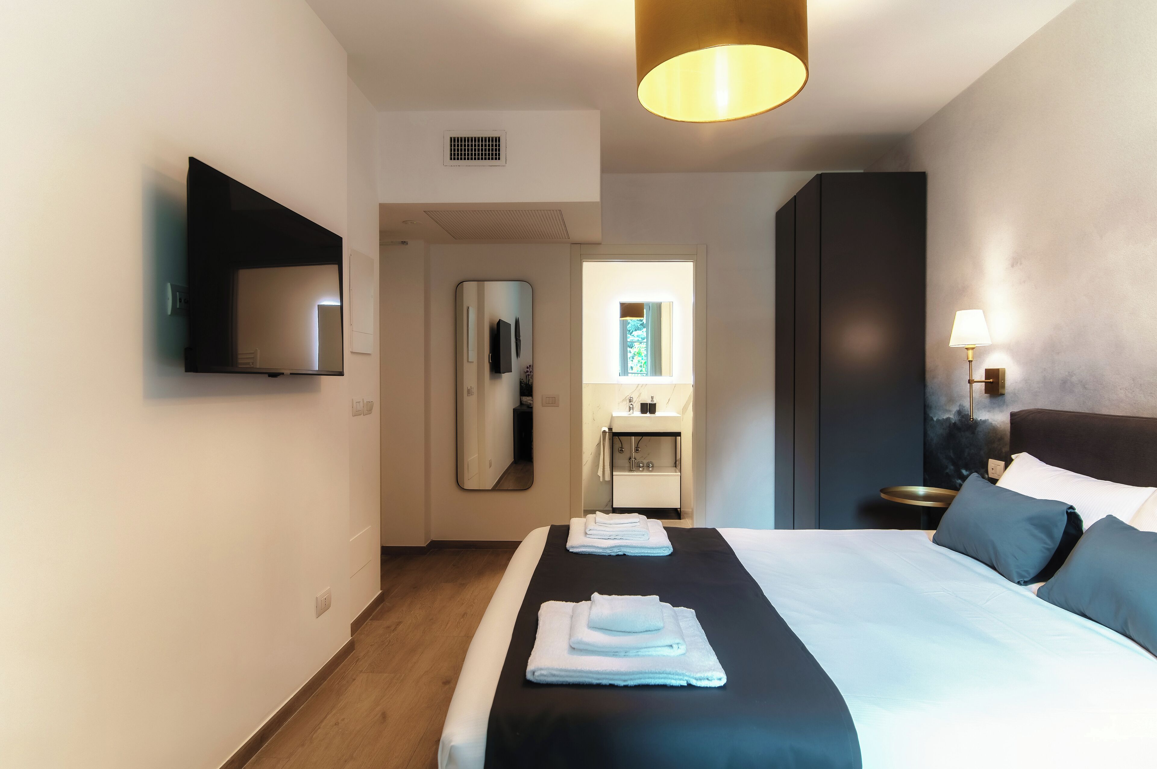 Habitación de hotel moderna con una cama grande, un televisor de pantalla plana montado en la pared, una mesita de noche con lámpara y vistas al baño a través de una puerta.