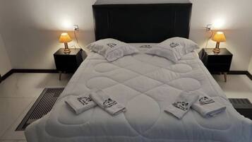 A neatly made bed with a white quilt and pillows, two bedside tables with lamps, and a vent on the floor.