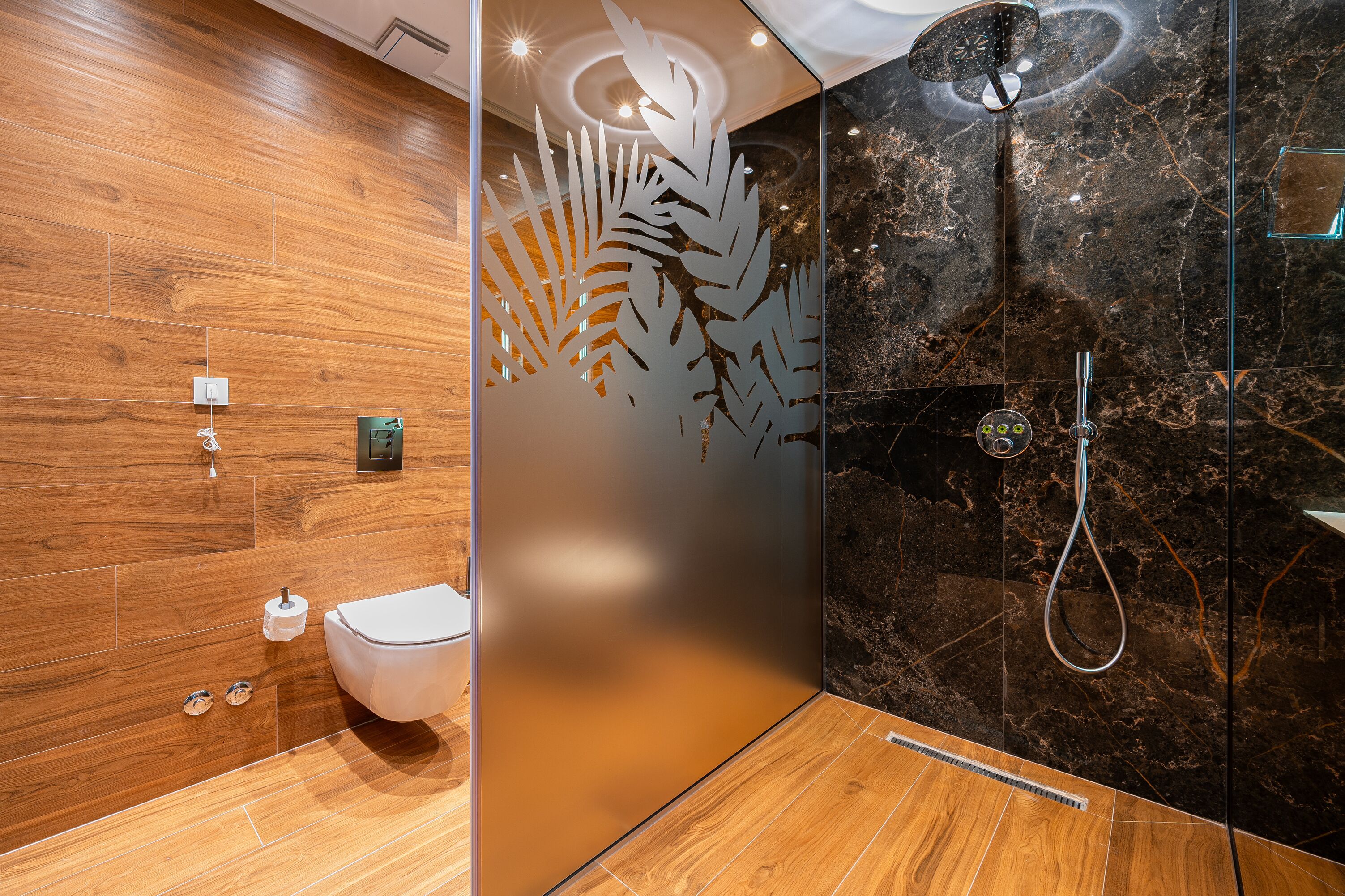 A modern bathroom with a wooden floor, a toilet, a shower with a glass door, and a decorative leaf pattern on the wall.