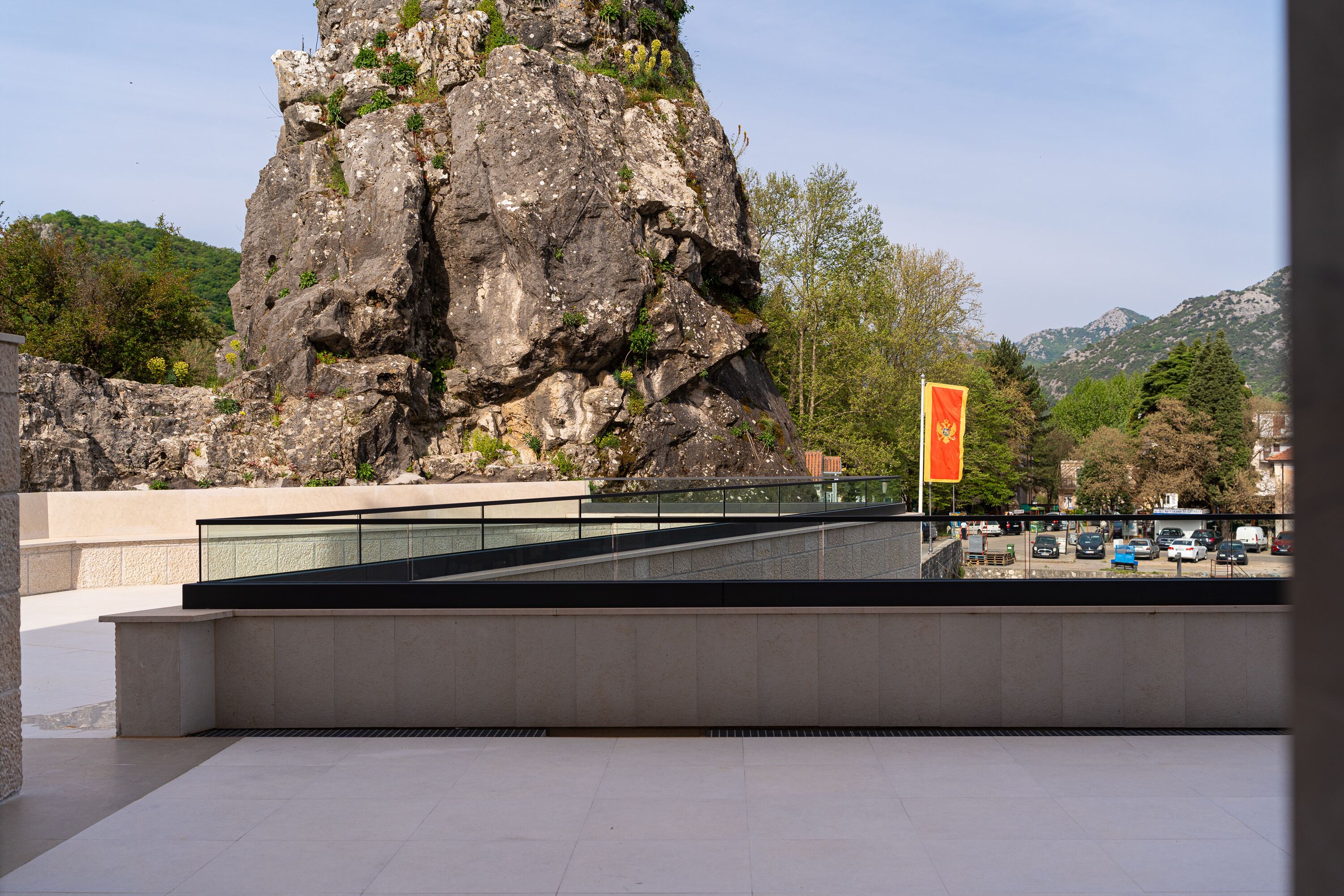 A large rock formation with a flag, a paved area, and a view of a parking lot and buildings in the background.