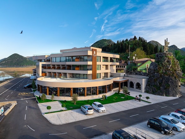A modern hotel with a curved facade, multiple balconies, and a parking area. - Hotel De Andros Virpazar (Virpazar)