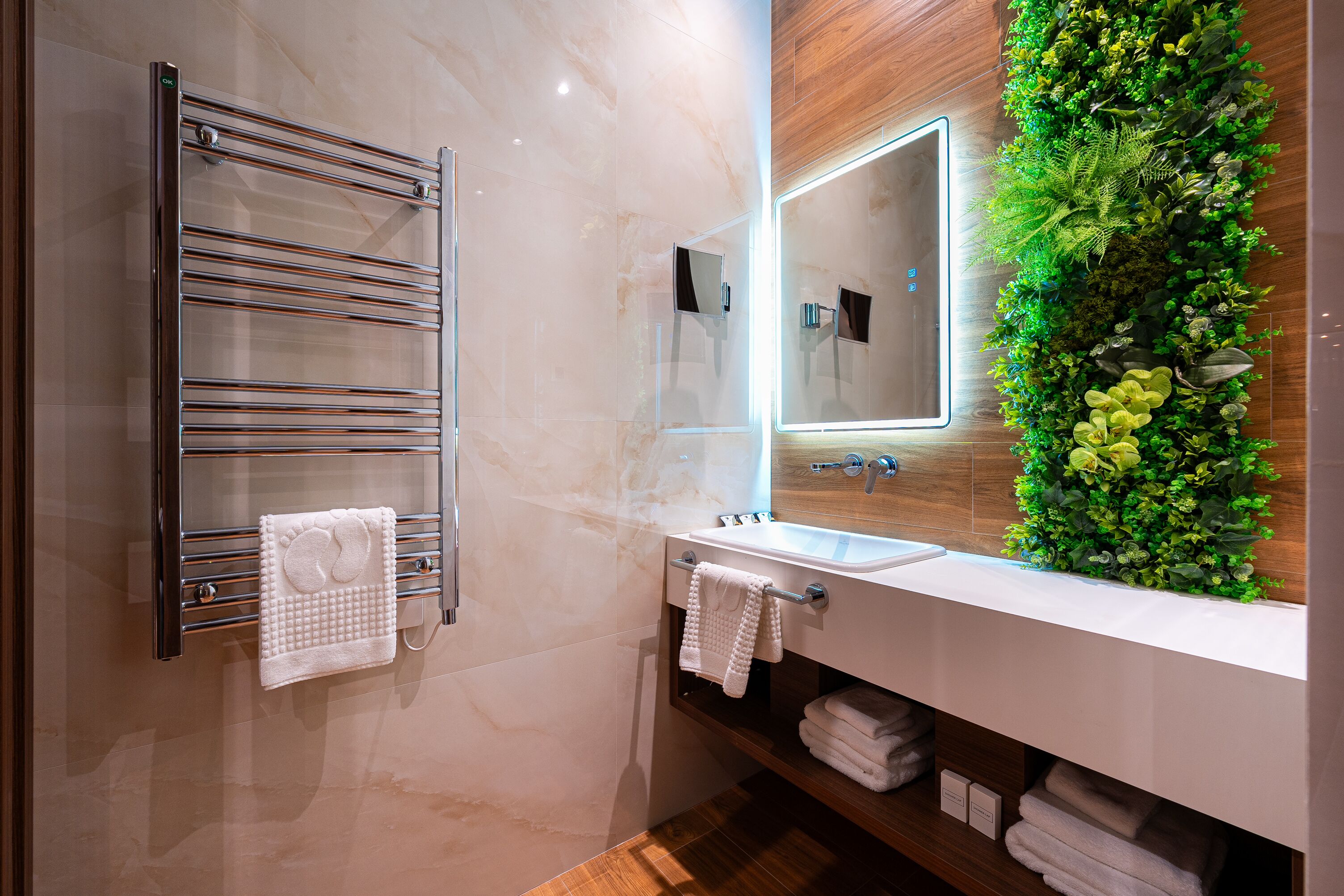 Un baño moderno con cabina de ducha de vidrio, toallero cromado, lavamanos blanco y espejo con jardín vertical verde.