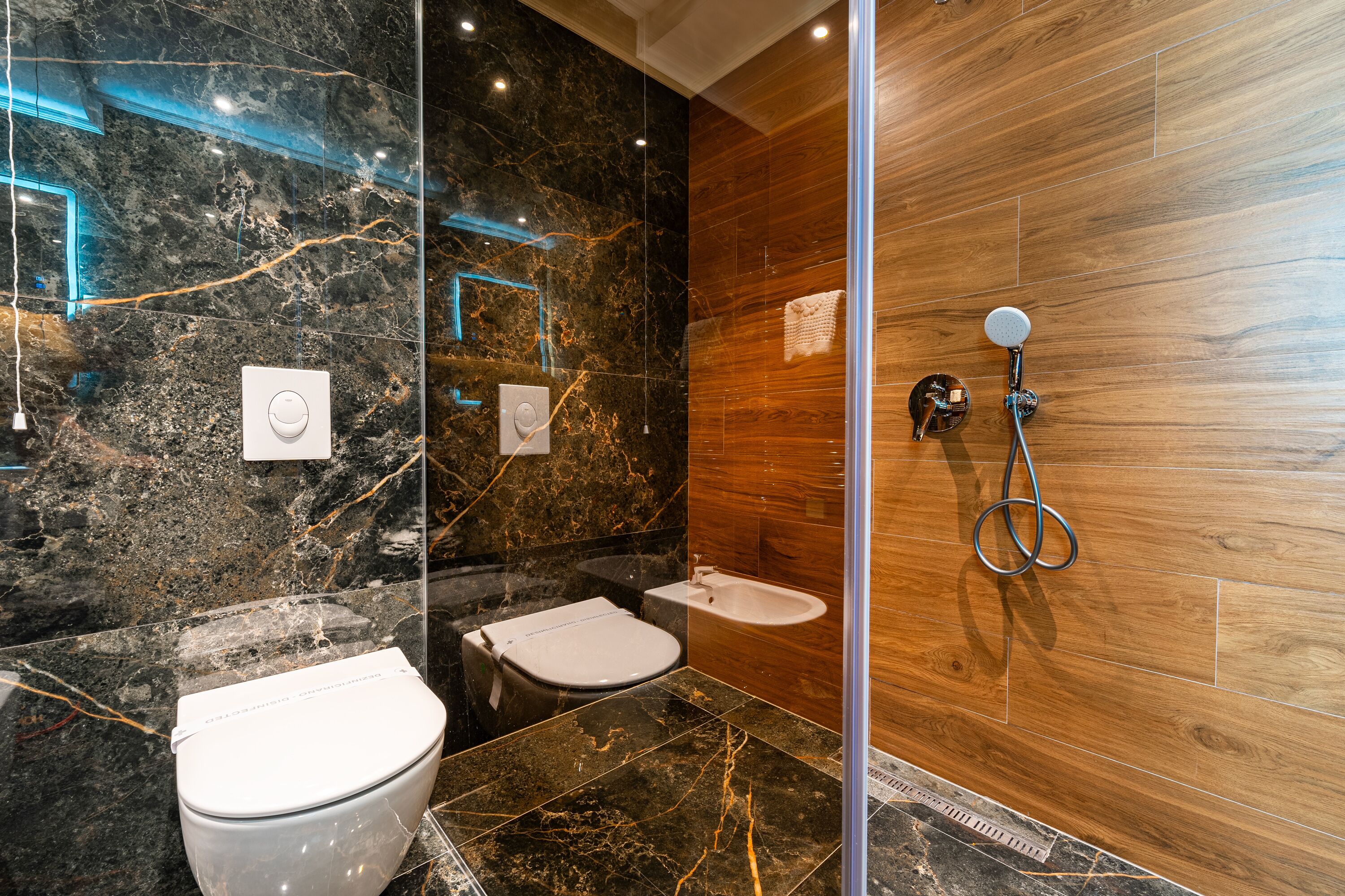 A modern bathroom with a marble vanity, a separate toilet, a bidet, and a shower with a wooden wall.