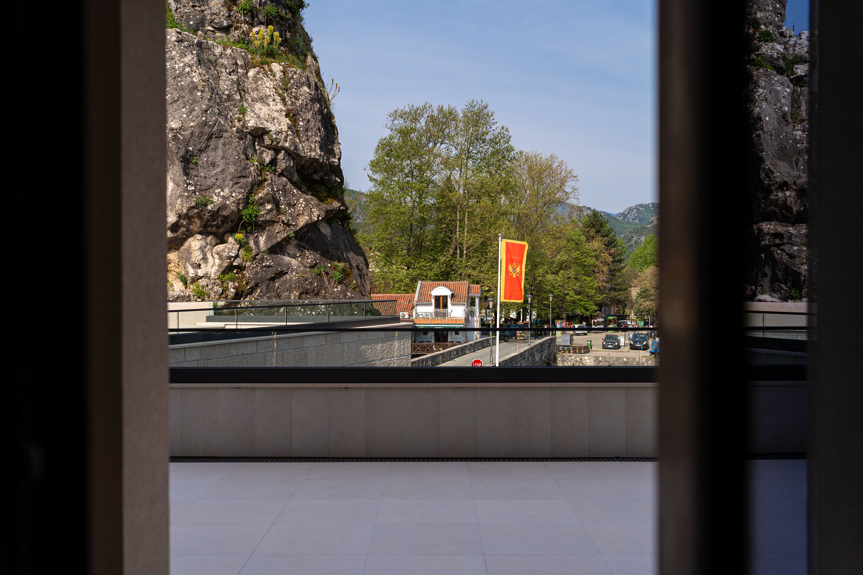 A view through a window of a building with a stone wall, a flag, and a distant building.