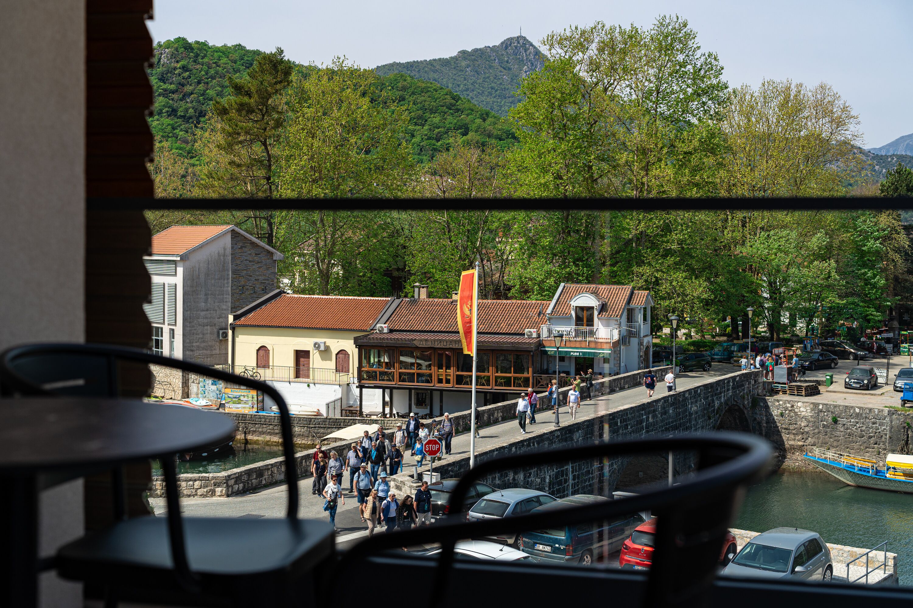 A view from a balcony overlooking a riverside walkway with people walking, a bridge, and buildings with stone facades.